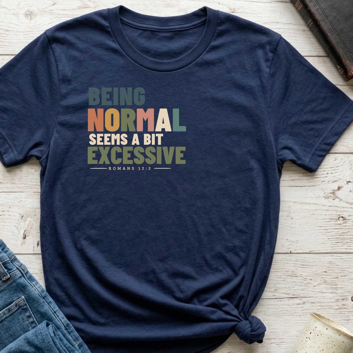 A navy blue graphic t-shirt laid flat on a light wood background, styled with a knot at the bottom hem and partially paired with blue jeans. The t-shirt features a retro-style text graphic reading 'BEING NORMAL SEEMS A BIT EXCESSIVE' with 'ROMANS 12:9' underneath.