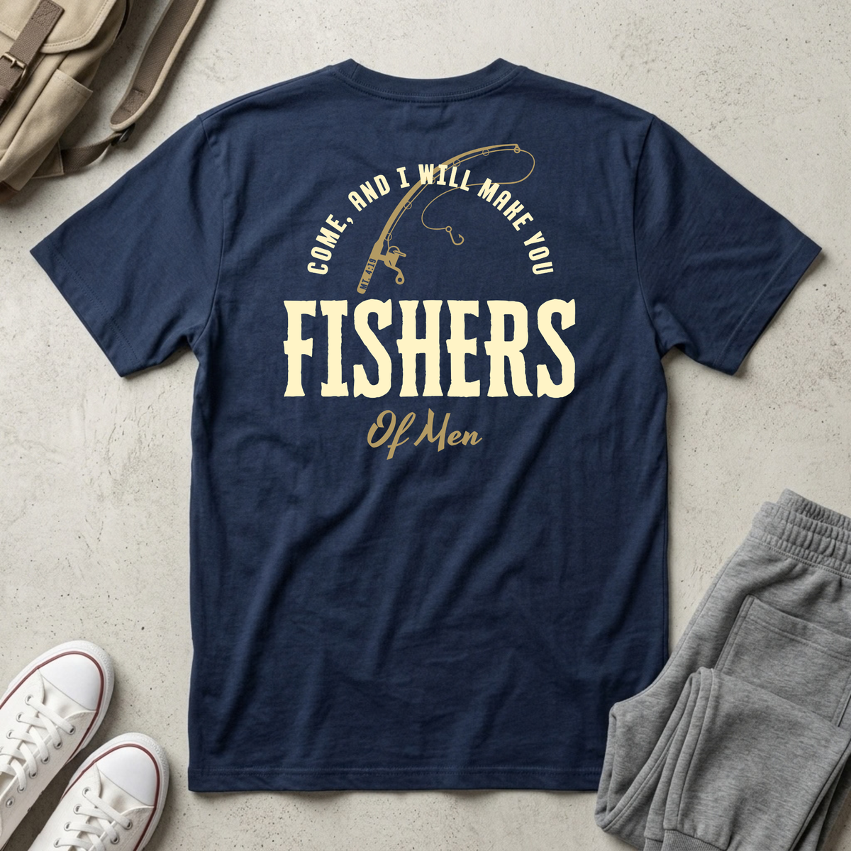 A navy blue short-sleeve Christian youth t-shirt with a vintage-style graphic on the back reading 'COME, AND I WILL MAKE YOU FISHERS OF MEN' in cream and gold lettering, styled in a flat-lay with gray sweatpants, white sneakers, and a tan backpack on a textured concrete floor.