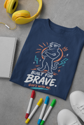 A navy blue Christian graphic T-shirt by SeedSowerGraphics laid flat on a gray surface, surrounded by yellow headphones, a notebook, three colored markers, and white sneakers. The shirt features a cartoon tiger wearing sunglasses with the text 'BUILT FOR BRAVE.