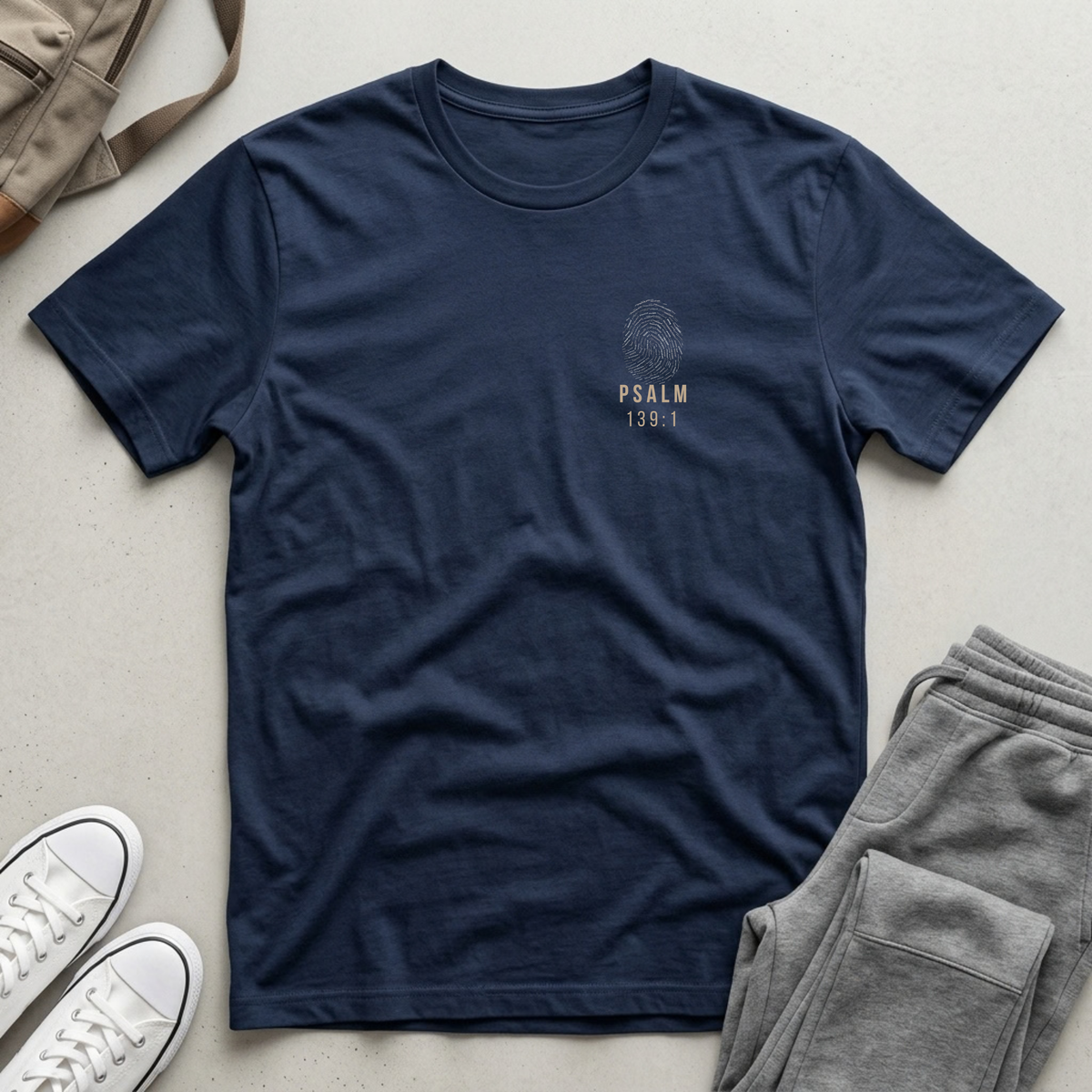A navy blue crewneck t-shirt with a small beige graphic on the left chest reading 'PSALM 139:1' beneath a fingerprint design, styled in a flat lay with gray sweatpants, white sneakers, and a tan backpack on a light gray surface.