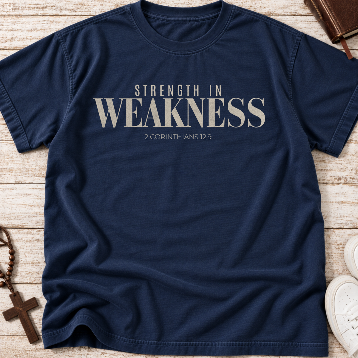 A navy blue short-sleeve t-shirt with the text 'STRENGTH IN WEAKNESS 2 CORINTHIANS 12:9' printed in beige on the chest, laid flat on a light wood background with decorative props.