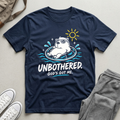 A navy blue Christian graphic t-shirt by SeedSowerGraphics laid flat on a light beige surface, styled with gray sweatpants, white sneakers, and a tan backpack. The t-shirt features a cartoon polar bear wearing sunglasses and floating in a ring with the text 'UNBOTHERED.