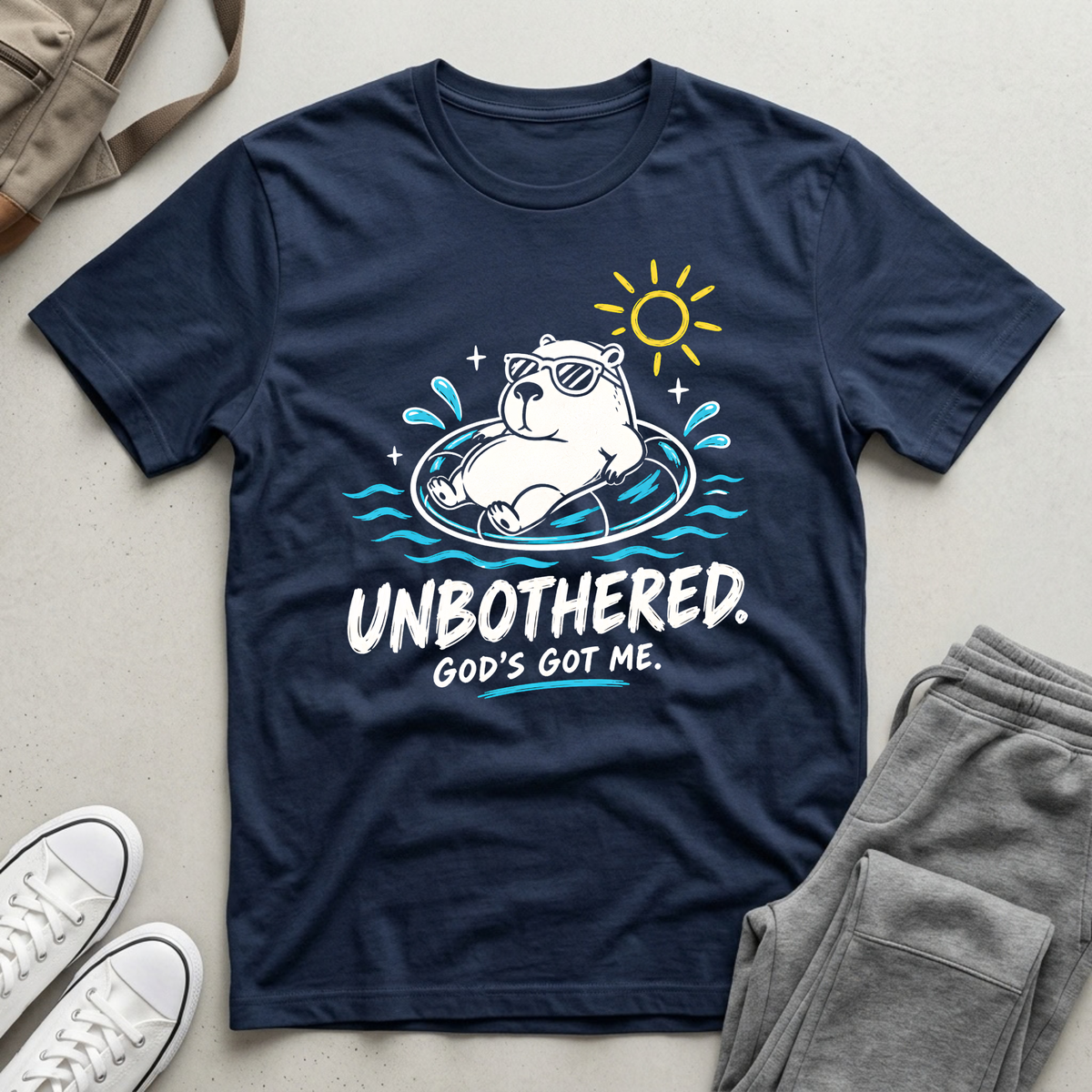 A navy blue Christian graphic t-shirt by SeedSowerGraphics laid flat on a light beige surface, styled with gray sweatpants, white sneakers, and a tan backpack. The t-shirt features a cartoon polar bear wearing sunglasses and floating in a ring with the text 'UNBOTHERED.