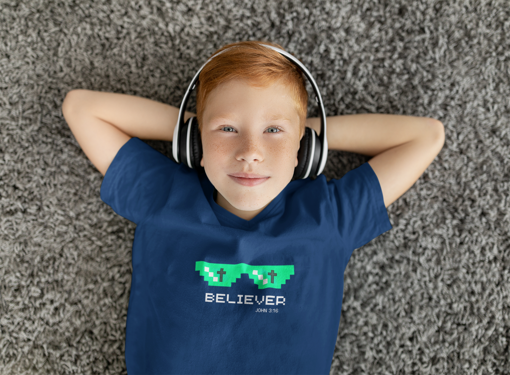 A young boy with red hair lies on a gray shag carpet wearing a navy blue Christian gaming t-shirt with a green pixelated glasses graphic and the word 'BELIEVER' printed on the chest. He is also wearing white over-ear headphones.