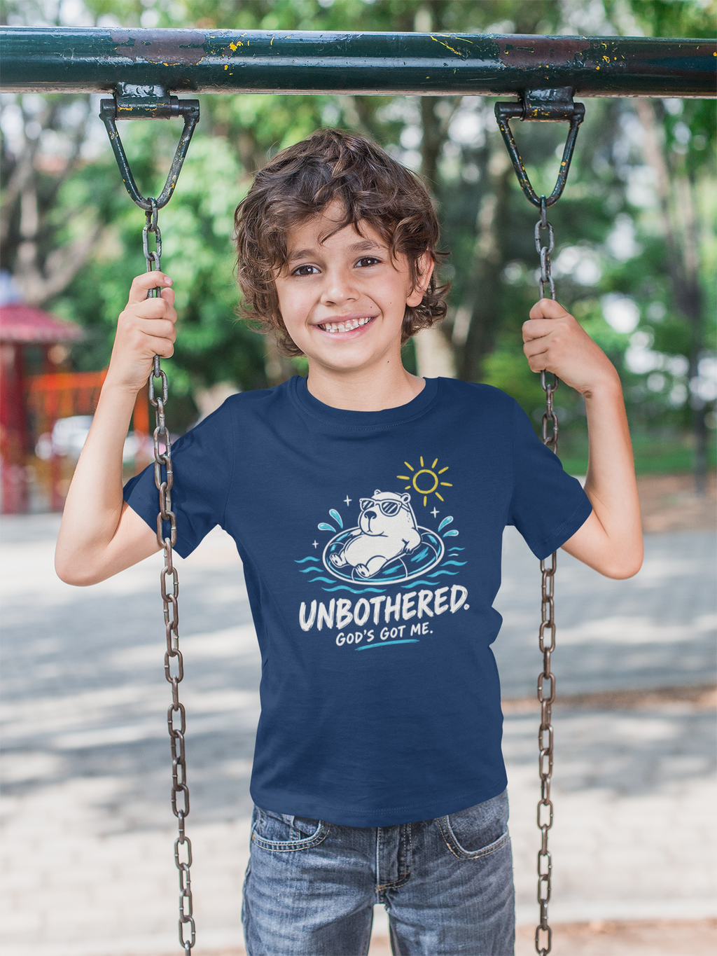 A smiling young boy stands on a playground swing holding the chains, wearing a navy blue Chrisian graphic t-shirt by SeedSowerGraphics with a cartoon bulldog and the text 'UNBOTHERED. GOD'S Got ME.