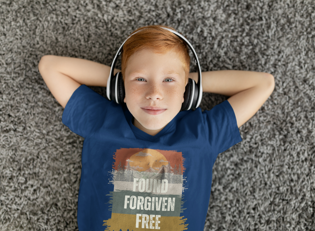 A young boy with red hair lies on a gray shag carpet, wearing over-ear headphones and a blue Chrisitan youth graphic t-shirt with the words 'FOUND FORGIVEN FREE' printed on the front.