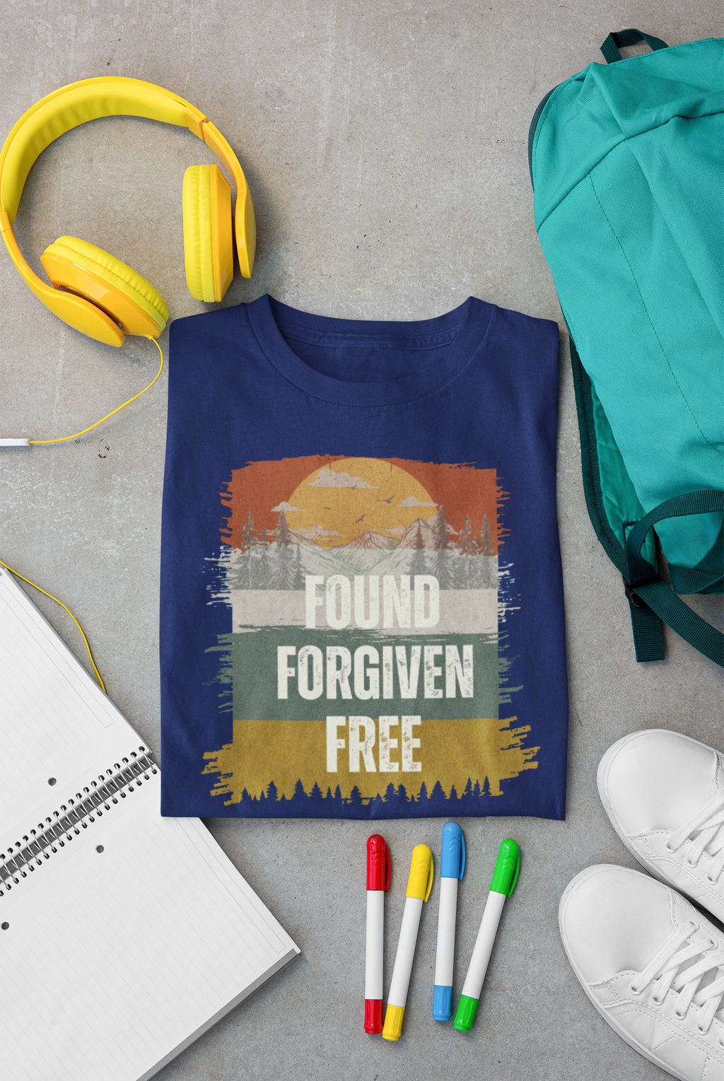 A navy blue Christian graphic t-shirt is laid flat on a gray concrete surface, surrounded by lifestyle props including yellow headphones, a teal backpack, a notebook, colored markers, and white sneakers. The t-shirt features a distressed sunset graphic with the words 'FOUND FORGIVEN FREE'.