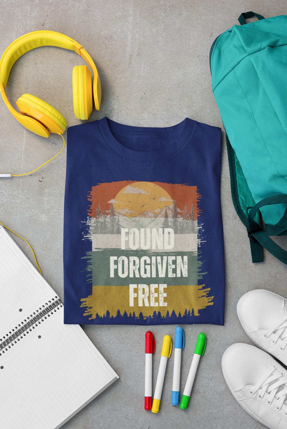 A navy blue Christian graphic t-shirt is laid flat on a gray concrete surface, surrounded by lifestyle props including yellow headphones, a teal backpack, a notebook, colored markers, and white sneakers. The t-shirt features a distressed sunset graphic with the words 'FOUND FORGIVEN FREE'.