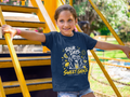 A young girl smiling while leaning on yellow outdoor stairs, wearing a navy blue Christian graphic T-shirt by SeedSowerGraphics with the text 'SOUR DAYS. SWEET GRACE.