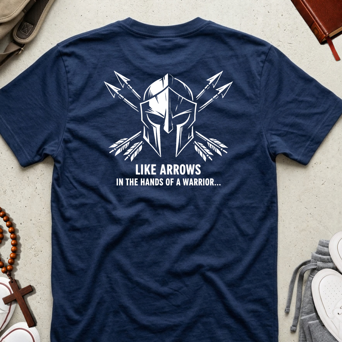 A navy blue short-sleeve t-shirt laid flat on a light textured surface, showing the back with a white graphic of a Spartan helmet and crossed arrows above the text 'LIKE ARROWS IN THE HANDS OF A WARRIOR.