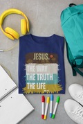 A navy blue graphic Christian t-shirt is laid flat on a gray concrete surface, surrounded by yellow headphones, a teal backpack, white sneakers, colored markers, and an open notebook. The t-shirt features a graphic with the text 'JESUS. The Way The Truth The Life."