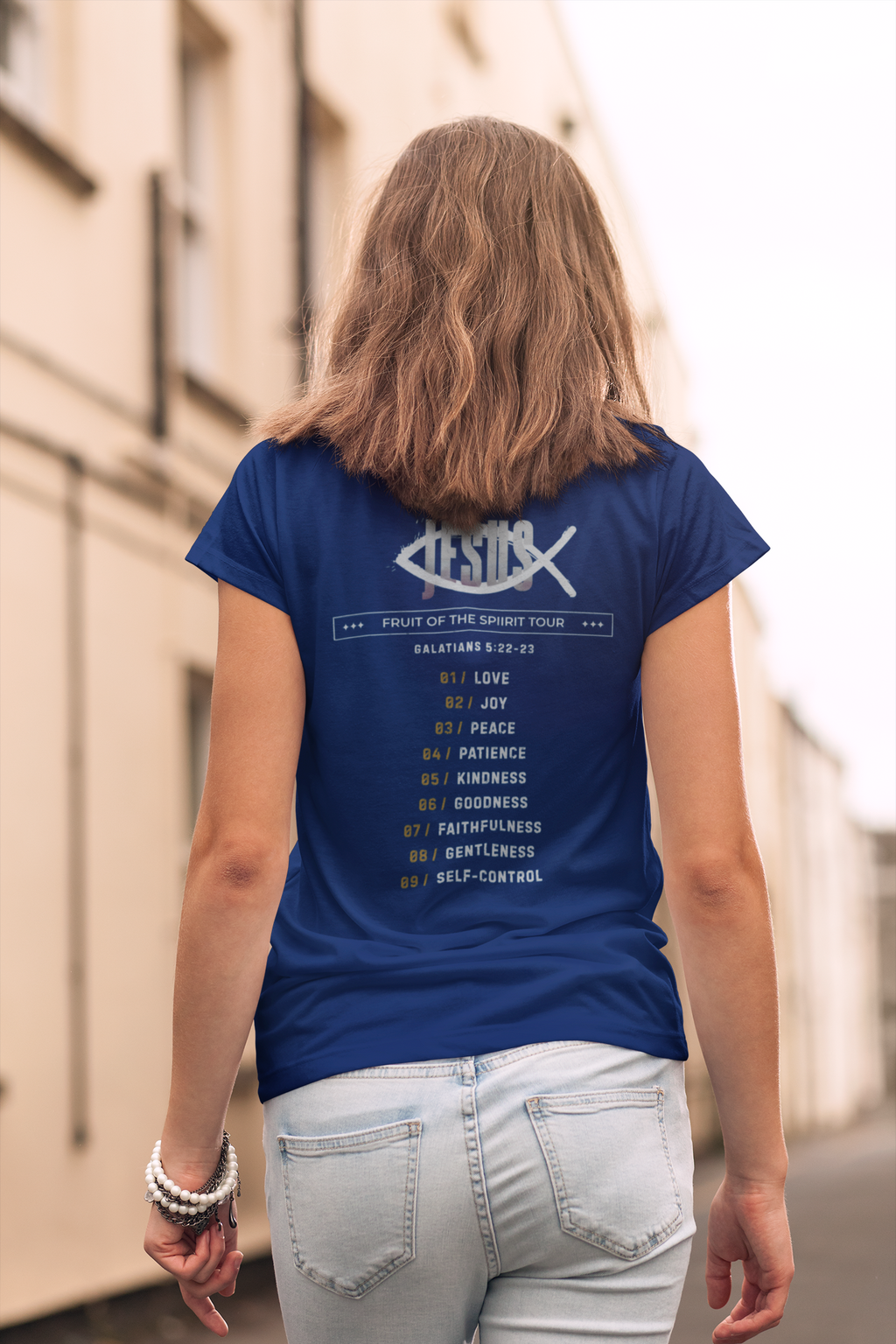 A woman viewed from behind wearing a navy blue Christian graphic t-shirt by SeedSowerGraphics with a white graphic print of the word 'Jesus' and a list of biblical virtues, paired with light-wash jeans in an outdoor street setting.