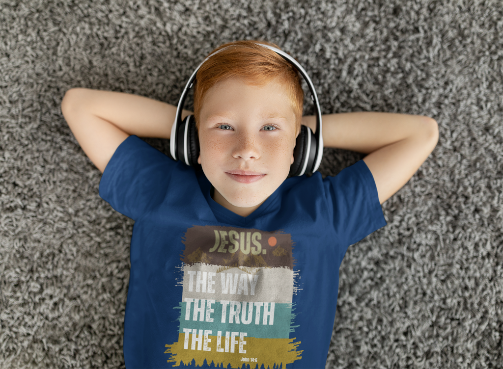 A young boy with red hair lies on a gray shag carpet, wearing a blue Christian graphic t-shirt and white over-ear headphones. The Chrisitan youth t-shirt features a distressed graphic with the text 'JESUS. The Way, The truth, the life."