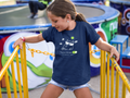 A young girl with her hair in a ponytail smiles while leaning on yellow metal railings at an outdoor event. She is wearing a navy blue Christianity t-shirt by SeedSowerGraphics with a white graphic of a cat wearing sunglasses and the text 'Peace Mode' with a green toggle switch.