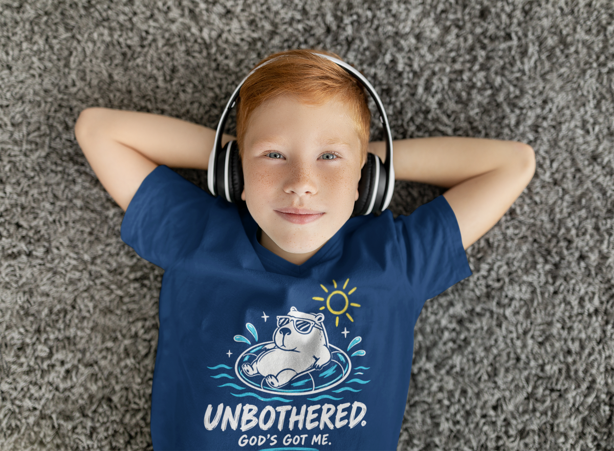 A red-haired boy lying on a gray shag carpet, wearing a navy blue Chrisitan graphic T-shirt by SeedSowerGraphics with a white capybara in a float and the text 'UNBOUGHTERED. GOD'S GOT ME.