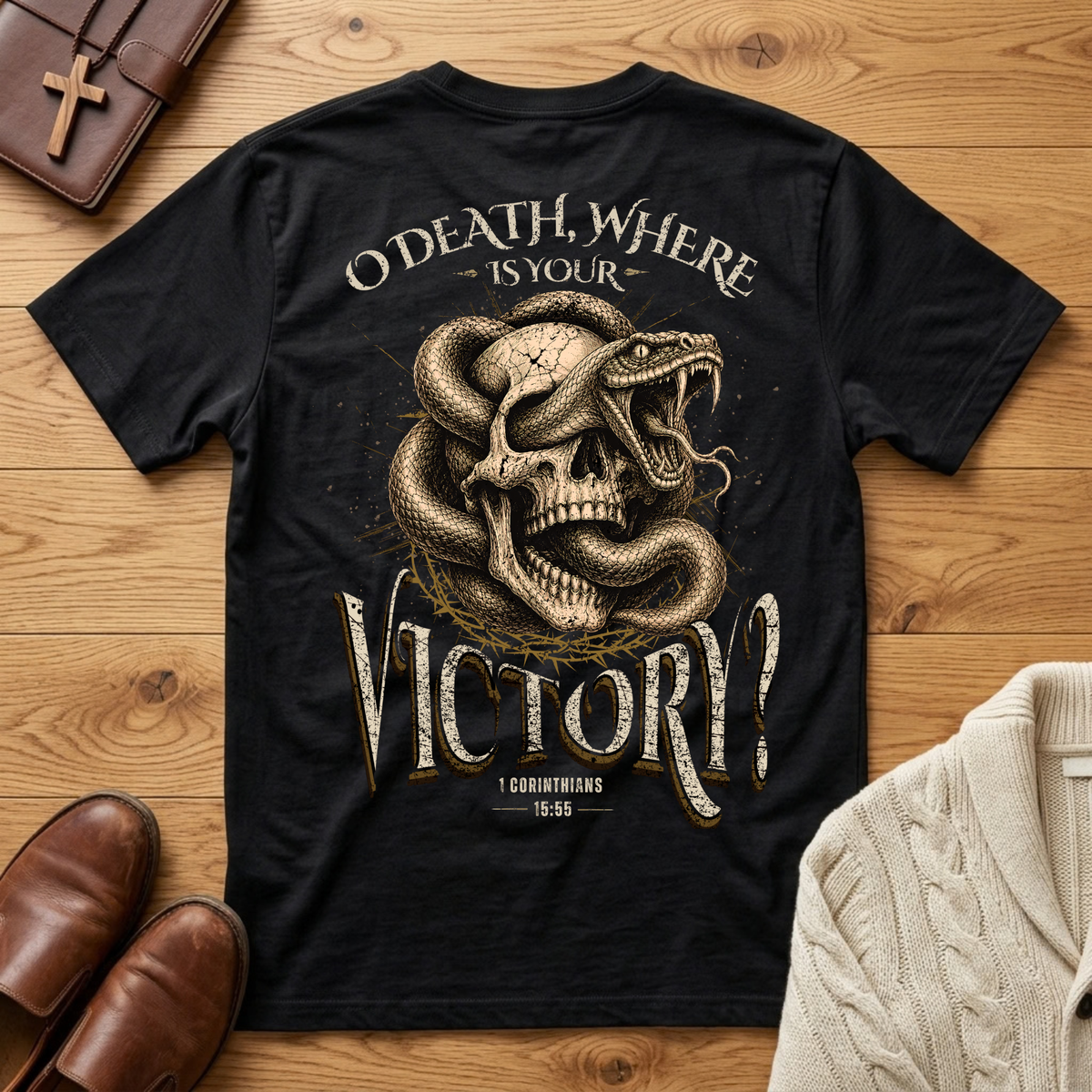 A black graphic t-shirt laid flat on a wooden surface, featuring a large design of a snake wrapped around a skull with the text 'O DEATH, WHERE IS YOUR VICTORY?'. The shirt is styled in a flat lay with brown leather shoes, a brown cross, a brown wallet, and a cream cable-knit sweater.