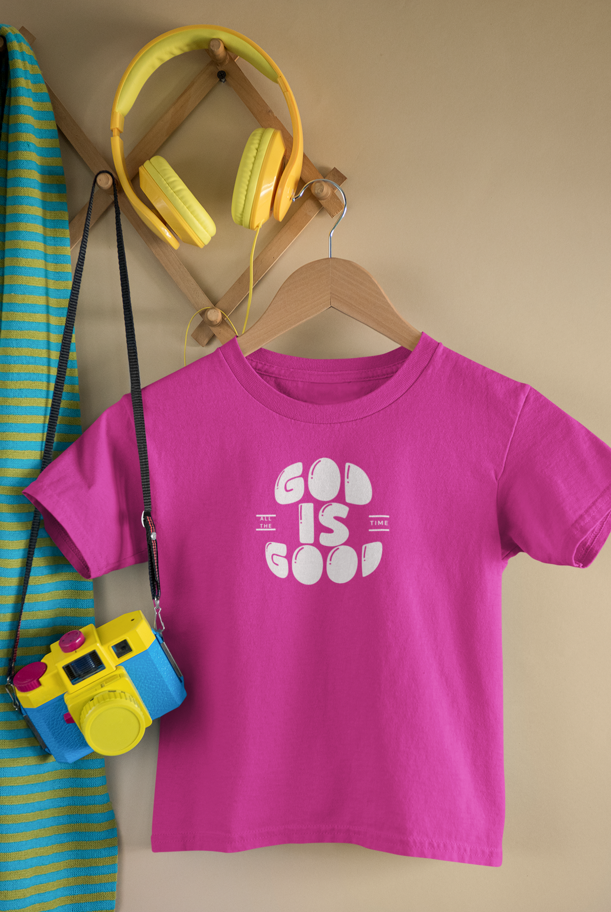 A bright pink short-sleeve Christian graphic t-shirt by SeedSowerGraphics with a white graphic reading 'GOD IS GOOD' hangs on a wooden hanger against a beige wall. Yellow over-ear headphones are on a wooden rack above, and a blue-and-yellow striped garment with a yellow camera accessory is partially visible on the left.