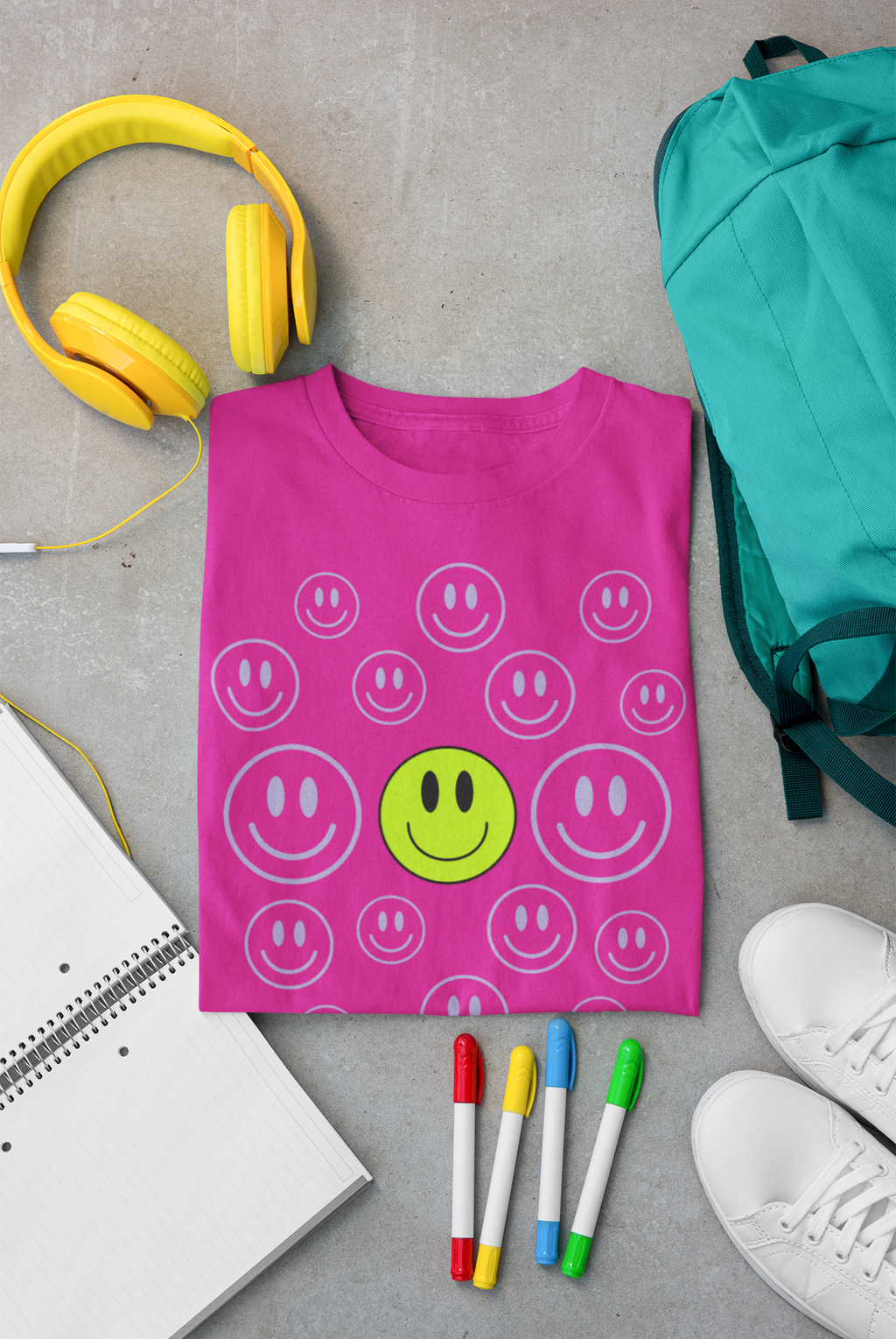 A folded bright pink Christian based T-shirt by SeedSowerGraphics  with a smiley face pattern is laid flat on a gray concrete surface, surrounded by school-style props including yellow headphones, a teal backpack, white sneakers, colored markers, and an open notebook.