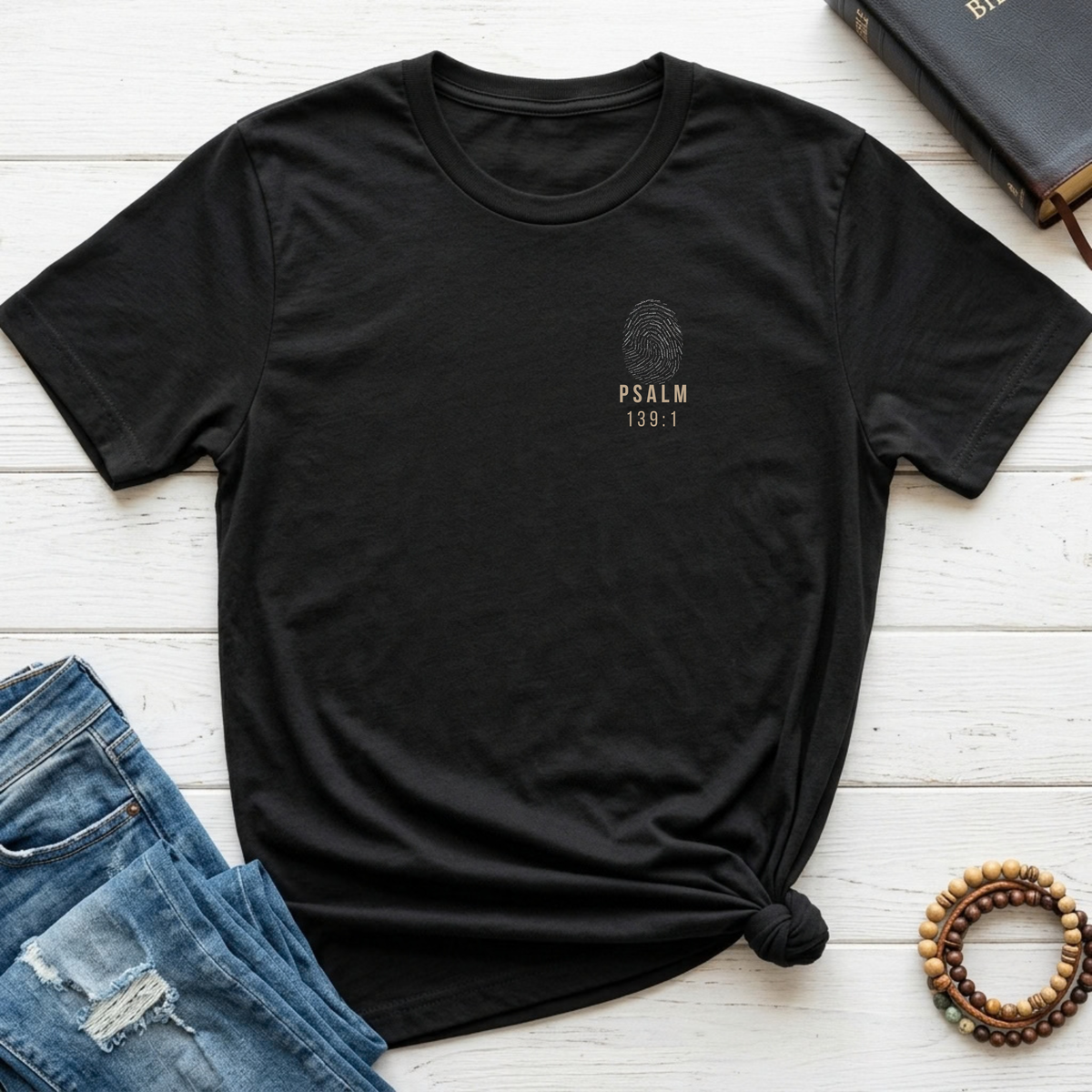 A black short-sleeve t-shirt with a small gold graphic on the left chest reading 'PSALM 139:1' with a fingerprint design, styled in a flat lay on white wooden planks with blue jeans, a book, and a wooden beaded bracelet.