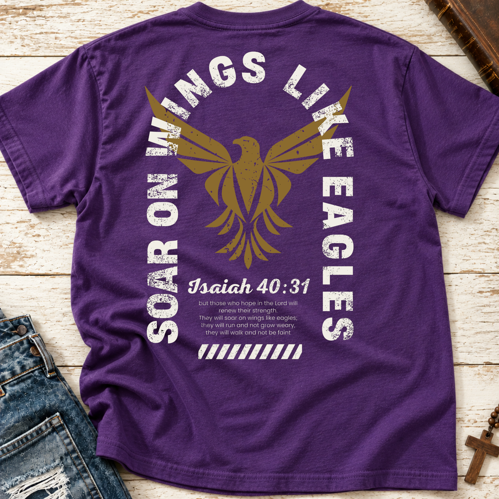 A purple short-sleeve t-shirt laid flat on a light wood surface, showing the back graphic with a large gold eagle, distressed white text reading 'Soar on Wings Like Eagles' and 'Isaiah 40:31', plus smaller scripture text. Jeans and part of a wooden necklace a Bible are visible as styling props.