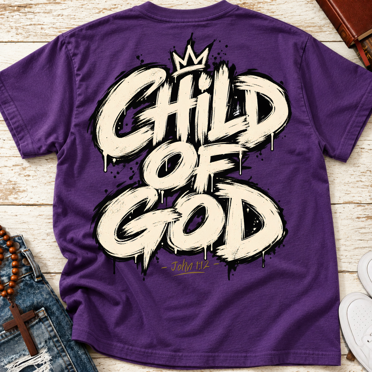 A purple Christian t-shirt laid flat on a gray whitewashed wooden background, featuring a large white and black spray-paint style graphic that reads 'CHILD OF GOD' with a small crown above and 'John 1:12' below. blue jeans are partially visible on the left, and a leather bound journal on the top right, and white sneakers  on the bottom right.