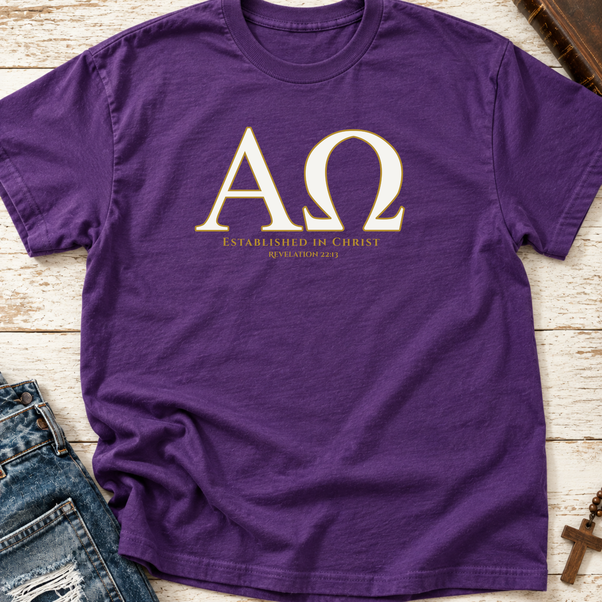 A purple short-sleeve t-shirt with a gold Greek letter design ('ΑΩ' and 'ESTABLISHED IN CHRIST') laid flat on a white wood background with jeans and a cross as props.