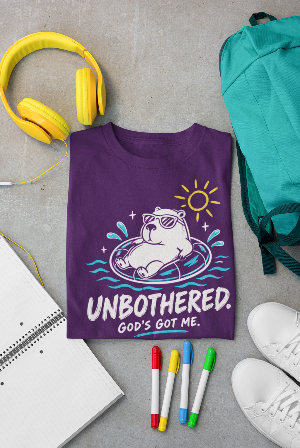 A purple Christian graphic T-shirt by SeedSowerGraphics laid flat on a gray surface, surrounded by yellow headphones, a teal backpack, a notebook, four colored markers, and white sneakers. The shirt features a cartoon capybara floating in a pool float with the text 'UNBOOTHERED.