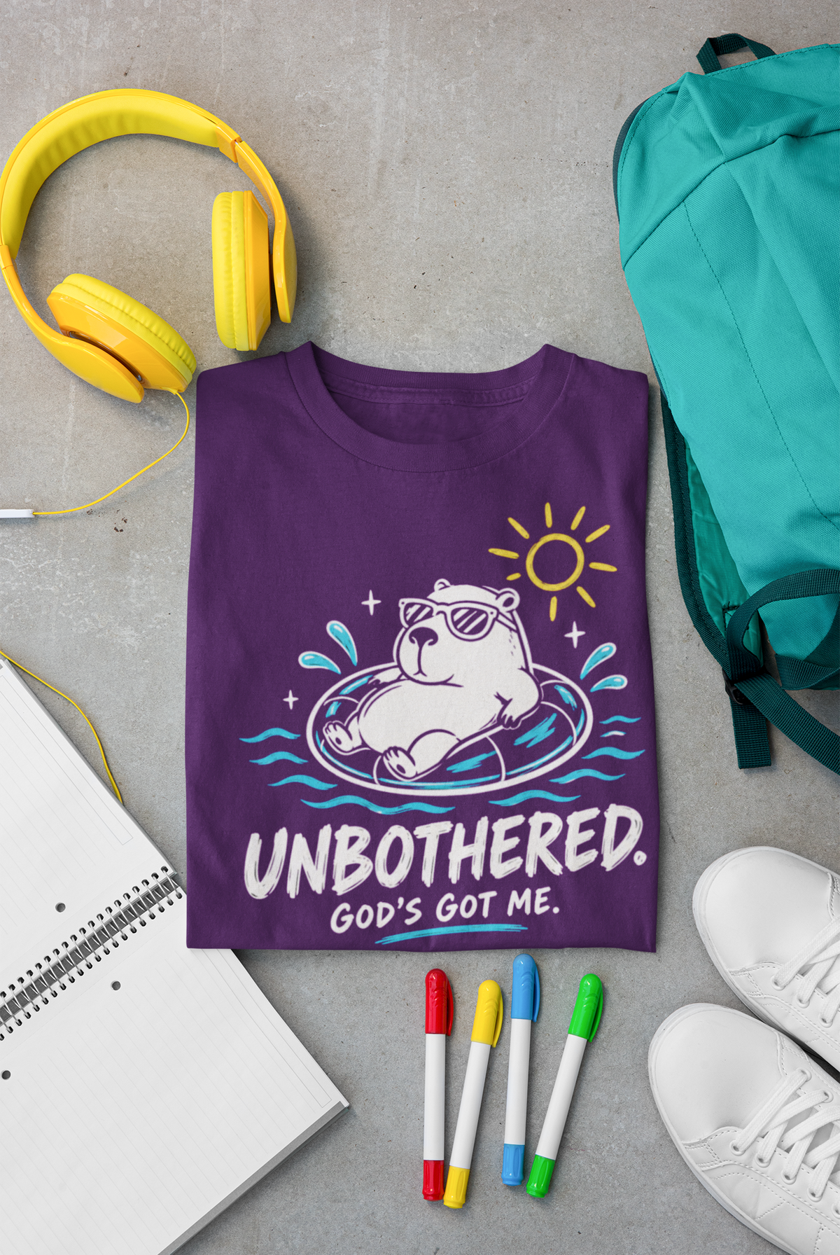 A purple Christian graphic T-shirt by SeedSowerGraphics laid flat on a gray surface, surrounded by yellow headphones, a teal backpack, a notebook, four colored markers, and white sneakers. The shirt features a cartoon capybara floating in a pool float with the text 'UNBOOTHERED.