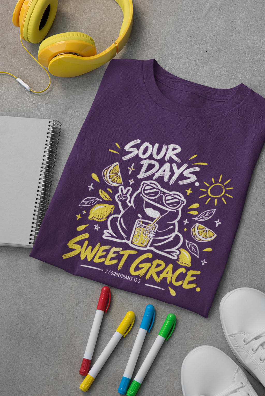 A purple Christian graphic T-shirt by SeedSowerGraphics laid flat on a gray surface, surrounded by yellow headphones, a notebook, colored markers, and white sneakers. The shirt features a frog wearing sunglasses and a drink, with the text 'SOUR DAYS, SWEET GRACE.