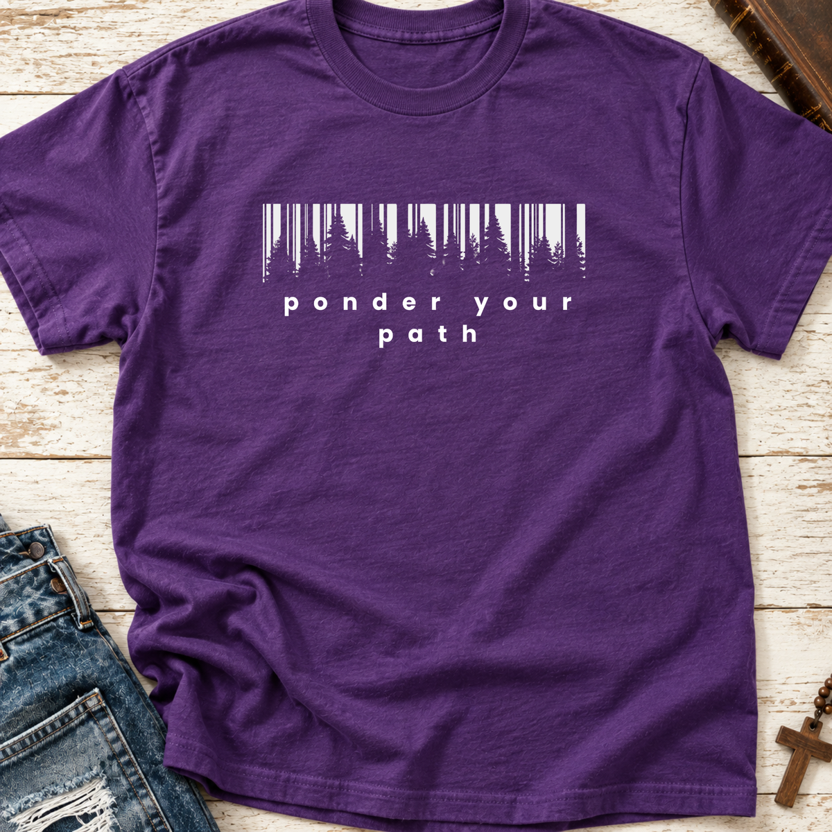 A purple crewneck t-shirt with a white graphic print reading 'ponder your path' beneath a stylized barcode made of tree silhouettes. The shirt is displayed in a flat-lay arrangement on a light wood background, styled with blue jeans on the left and a brown leather book and cross on the right.