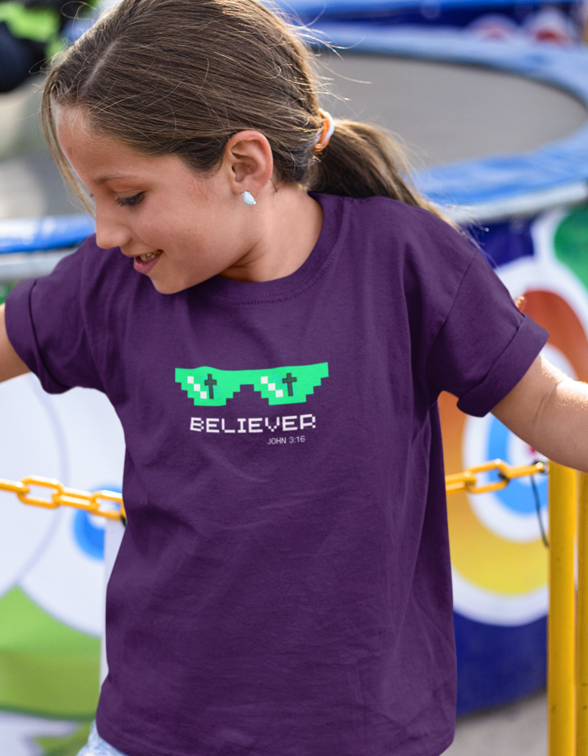 A young girl with her hair in a ponytail wearing a purple Christian gaming t-shirt with a green pixelated sunglasses graphic and the word 'BELIEVER' printed on the front, set against a colorful outdoor background.
