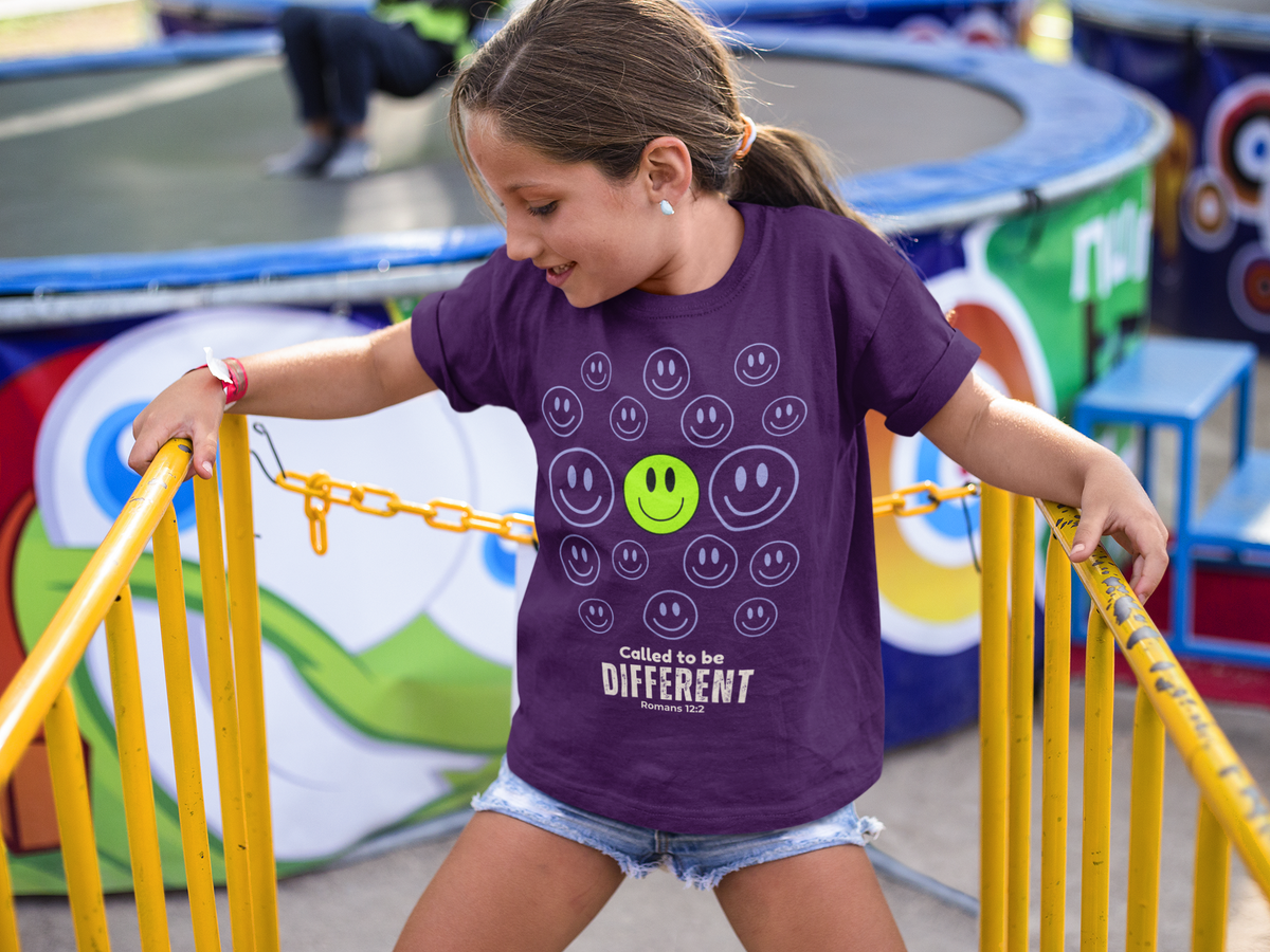 A young girl smiling while climbing a yellow metal gate at an outdoor fair, wearing a purple graphic t-shirt with smiley faces and the text 'Called to be DIFFERENT'.