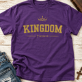 A purple short-sleeve crewneck t-shirt with a gold graphic print reading 'KINGDOM' under a crown, with 'MATTHEW 6:33' and a cross below. The shirt is laid flat on a white wood background with jeans and a cross necklace partially visible as props.
