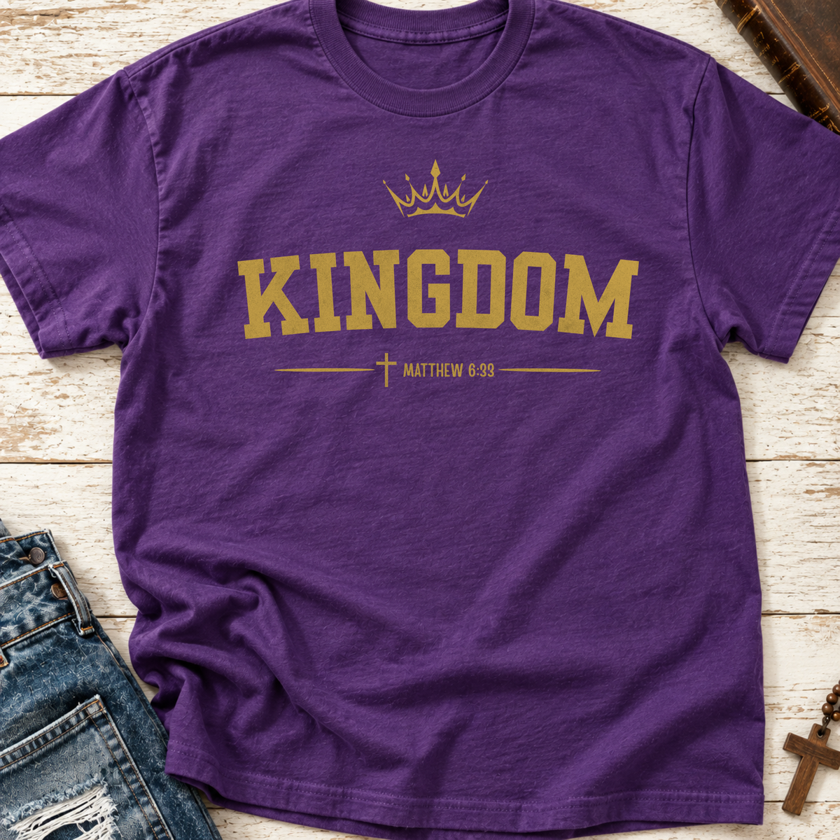 A purple short-sleeve crewneck t-shirt with a gold graphic print reading 'KINGDOM' under a crown, with 'MATTHEW 6:33' and a cross below. The shirt is laid flat on a white wood background with jeans and a cross necklace partially visible as props.