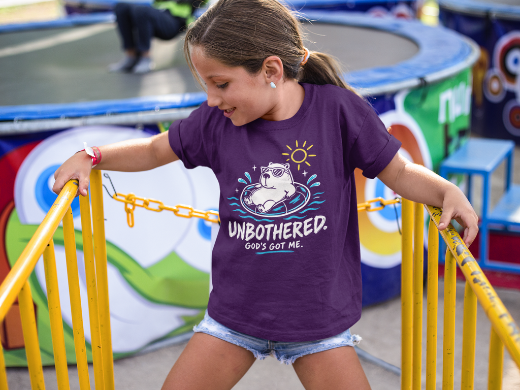 A young girl wearing a purple Christian graphic t-shirt by SeedSowerGraphics with the text 'UNBOTHERED GOD'S GOT ME' and a cartoon dog in a pool, posing at a playground with yellow railings.