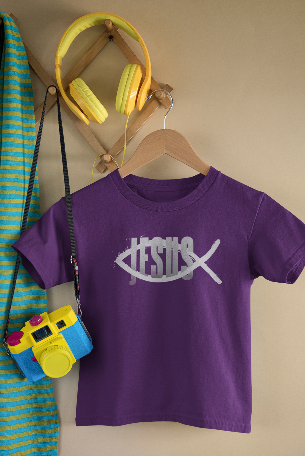 A purple short-sleeve Christian graphic t-shirt  by SeedSowerGraphics on a wooden hanger, featuring a white fish-shaped graphic with the word 'JESUS' across it. The shirt is styled in a room setting with a wooden wall rack holding yellow headphones, a blue and yellow striped towel, and a toy camera.