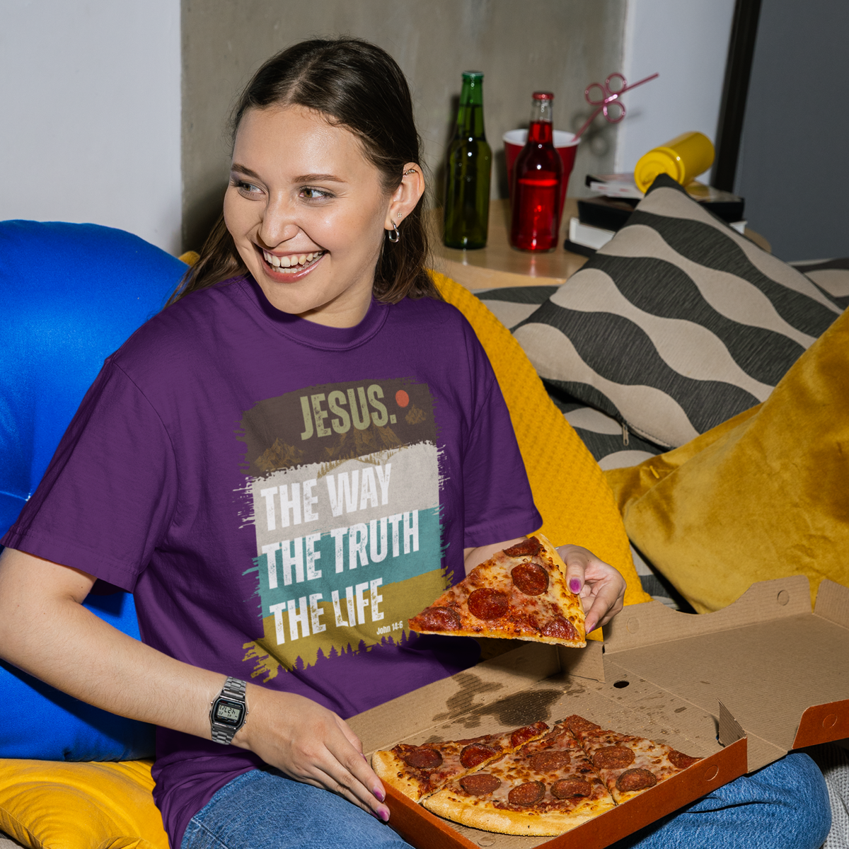 A woman sitting on a couch in a living room, wearing a purple graphic t-shirt with the text 'JESUS THE WAY THE TRUTH THE LIFE' and holding a slice of pizza next to an open pizza box.