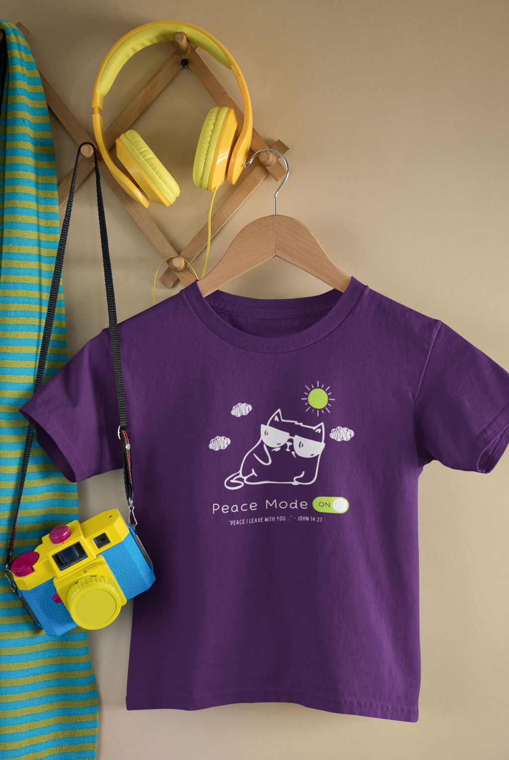 A purple short-sleeve Chrsitian graphic kids' t-shirt by SeedSowerGraphics with a cartoon cat wearing sunglasses and the text 'Peace Mode ON' hanging on a wooden hanger. The shirt is styled in a cozy room setting with a wooden wall rack holding yellow headphones, a blue and yellow camera, and a striped towel.