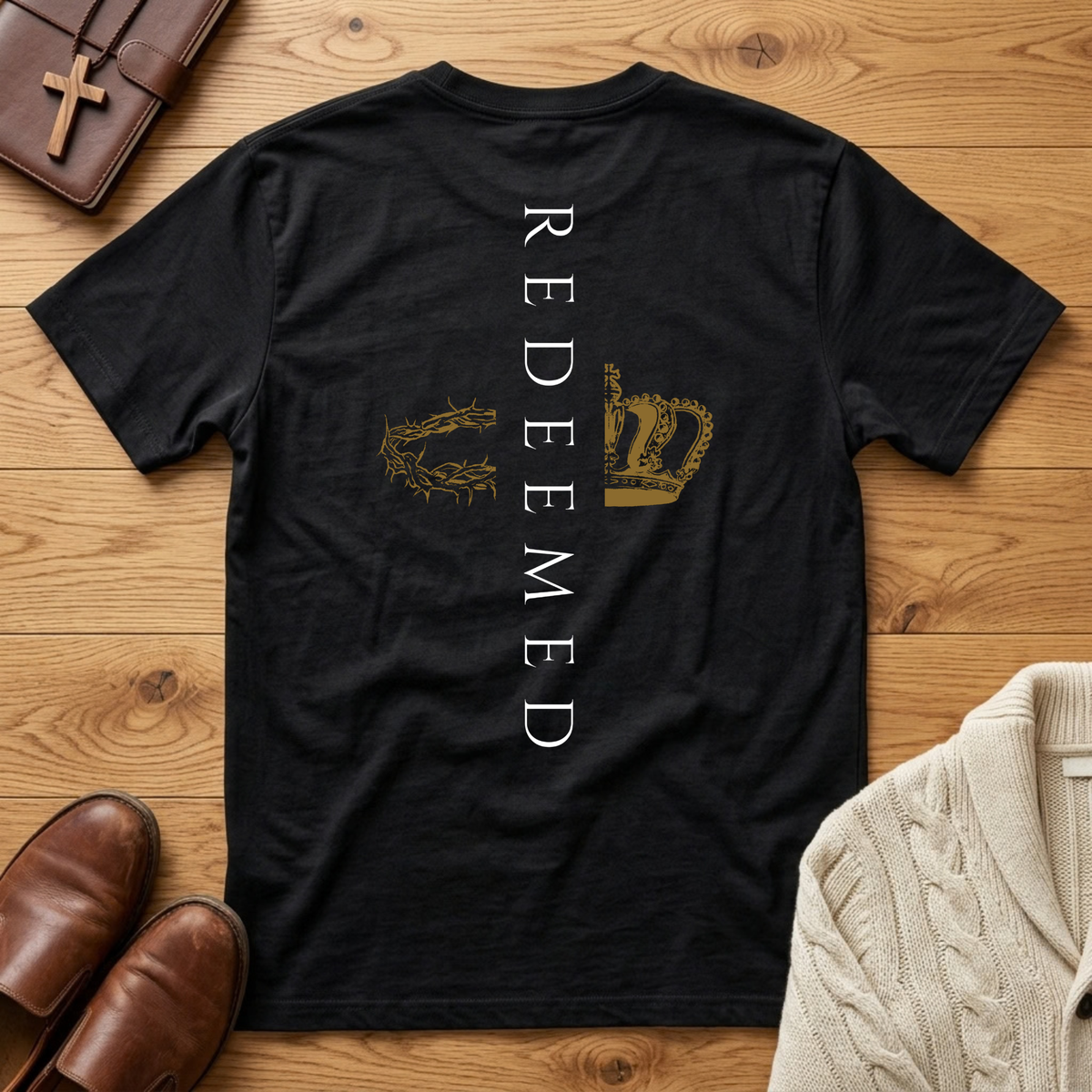 A black short-sleeve t-shirt laid flat on a wooden surface, featuring the word 'REDEEMED' printed vertically in white on the back, with a crown of thorns graphic on the left and a gold crown graphic on the right. Styled flat-lay props include brown leather loafers, a brown cross necklace, a cream cable-knit sweater, and a brown leather wallet.