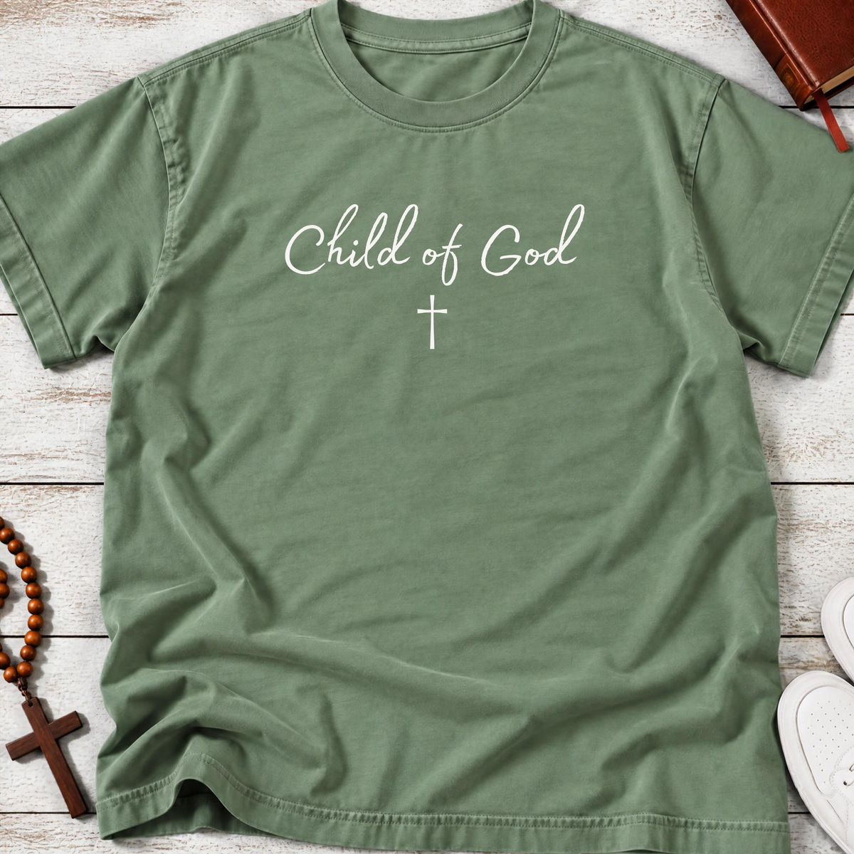 A sage green Christian short-sleeve t-shirt with 'Child of God' and a cross printed in white on the chest, laid flat on a light wood background with a rosary, book, and sneakers as props.