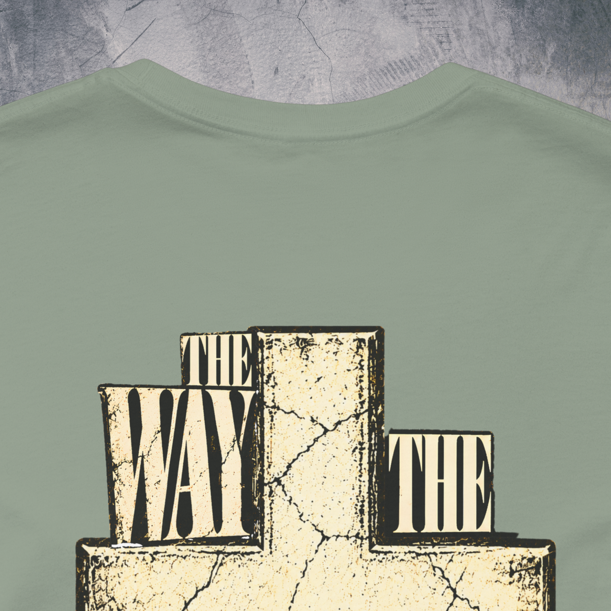 A close-up of a sage green Christian t-shirt by SeedSowerGraphics  featuring a graphic print of a cracked beige cross with the words 'THE WAY TO THE' in black and cream lettering.