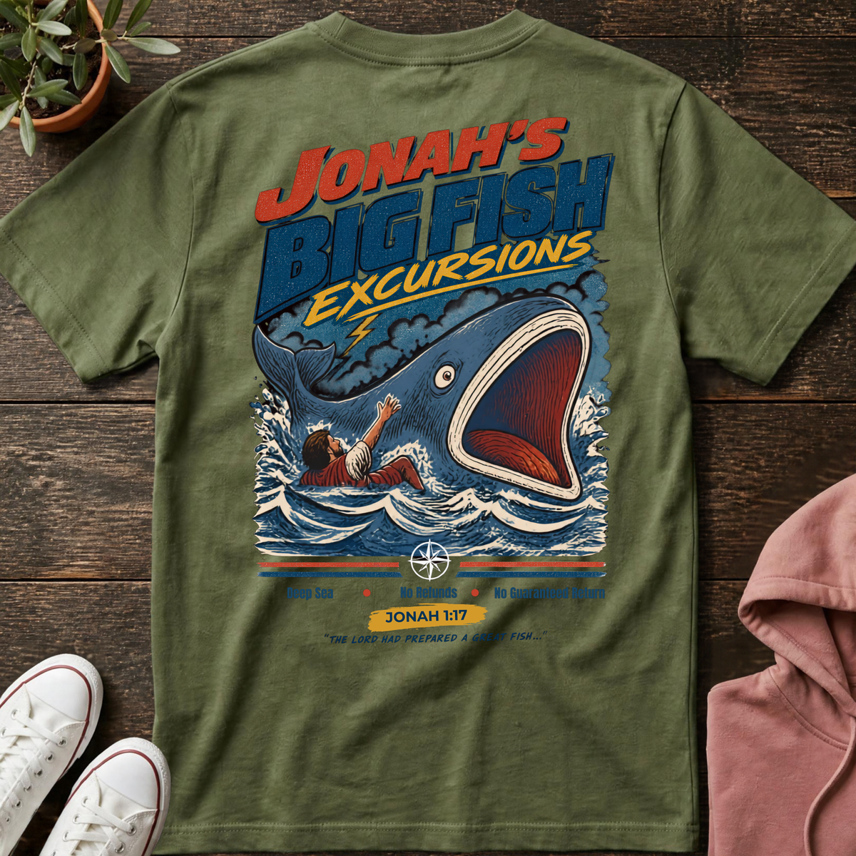An olive green graphic t-shirt laid flat on a dark wood background, styled with white sneakers, a pink hoodie, and a small potted plant. The t-shirt features a large retro-style graphic of a giant blue fish with its mouth open, swallowing a man in the water, with the text 'JONAH'S BIG FISH EXCURSIONS'.
