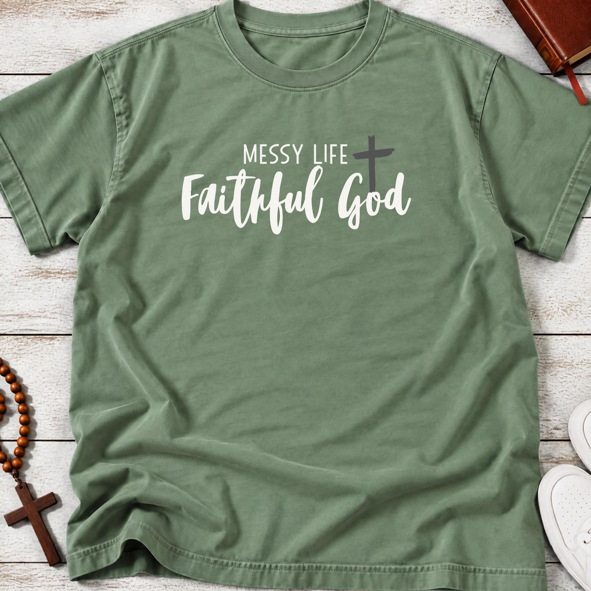 An olive green short-sleeve t-shirt with the text 'MESSY LIFE Faithful God' and a cross graphic printed on the chest, styled in a flat-lay on a white wood background with a rosary, book, and sneakers as props.