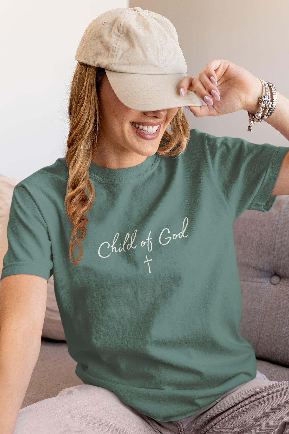 A woman wearing a sage green Christian faith t-shirt by SeedSowerGraphics with 'Child of God' and a cross printed in white on the chest, paired with a beige baseball cap and light gray pants, sitting on a gray couch.
