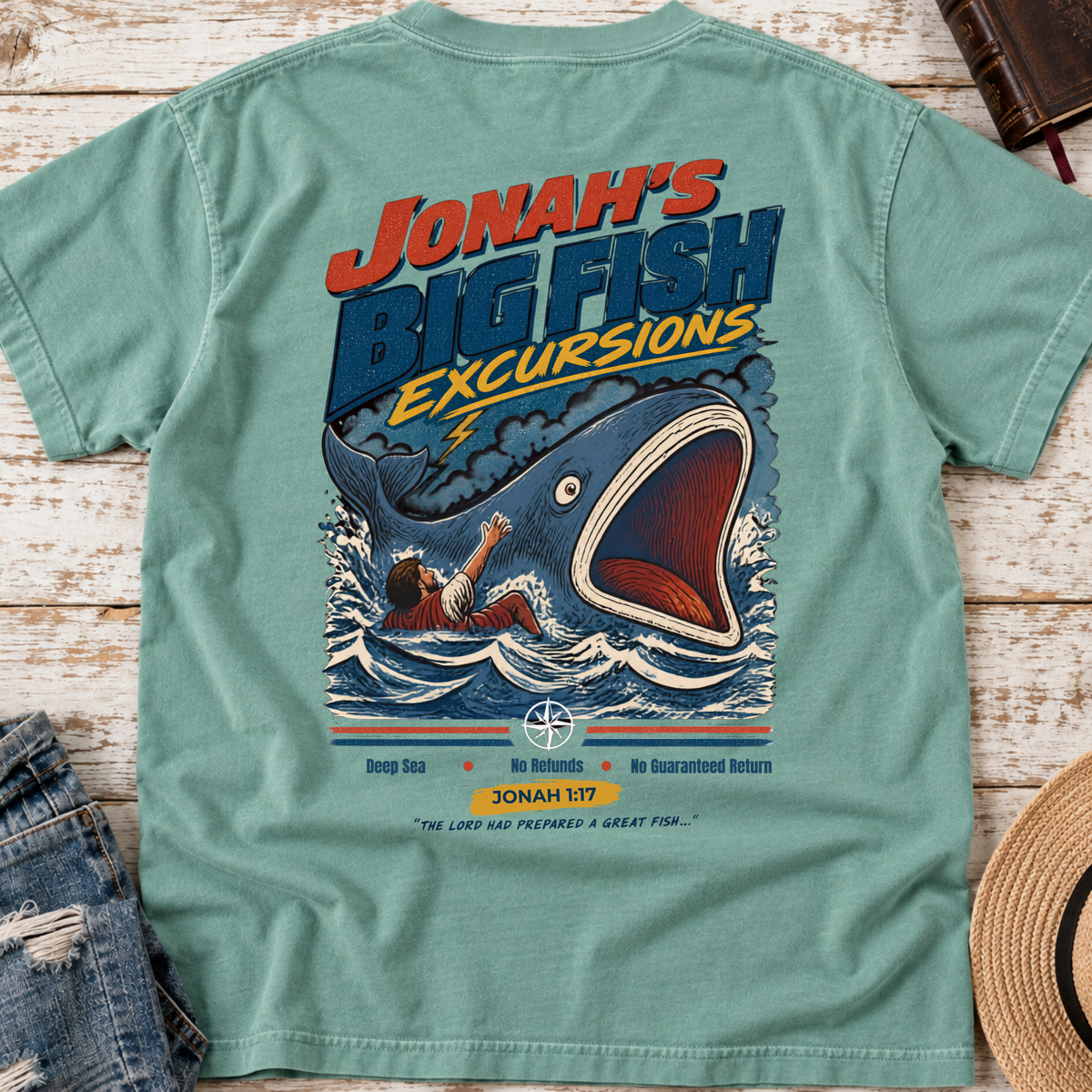 A seafoam green graphic t-shirt laid flat on a whitewashed wooden background, styled with a pair of distressed blue jeans on the left and a straw hat on the right. The back of the t-shirt features a large graphic print of a giant blue fish with its mouth open, catching a person in the water, with the text 'JONAH'S BIG FISH EXCURSIONS'.