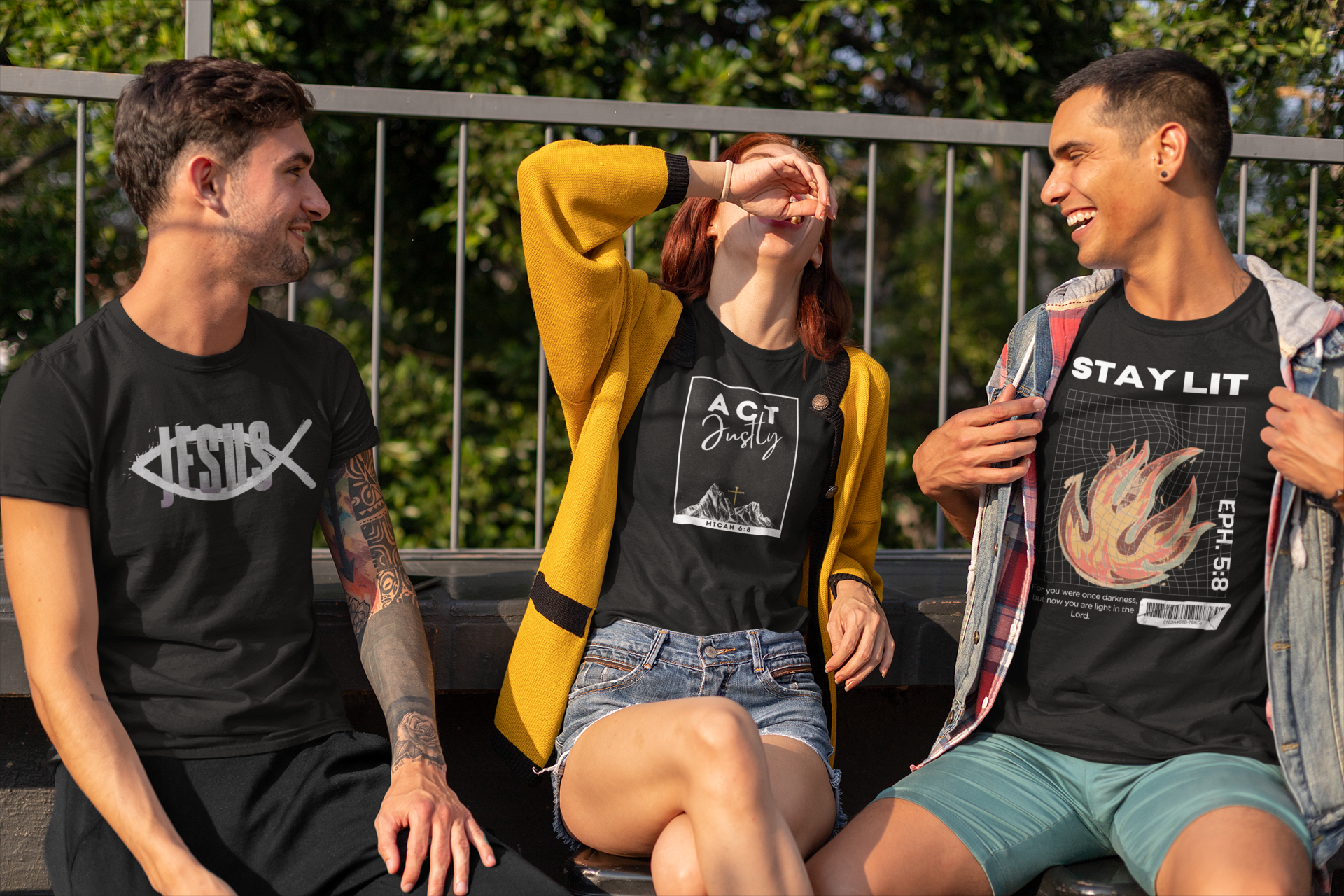 Three young adults sitting outdoors on a bench wearing black Christian graphic t-shirts by SeedSowerGraphics. The shirt on the left has a white fish logo with 'Jesus' text, the center shirt has a white graphic with 'Act Justly' text, and the right shirt has a flame graphic with 'STAY LIT' text.