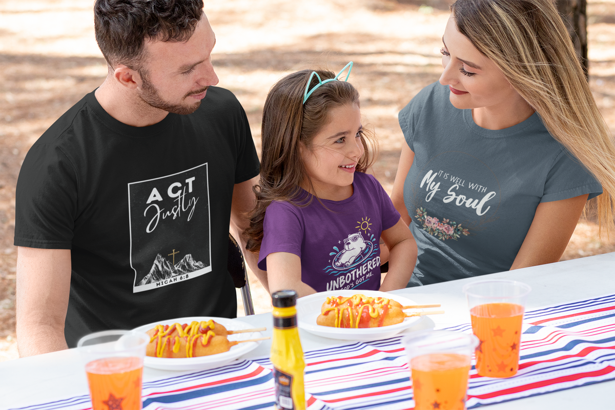A family of three wearing custom Christian graphic t-shirts sits at a table outdoors. The man on the left wears a black Christian t-shirt with a white graphic reading 'ACT Justly'.