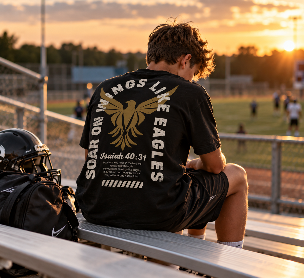 A young man sits on metal bleachers at a football field at sunset, wearing a black Christian t-shirt by SeedSowerGraphics with a large gold eagle graphic and the text 'SOAR ON WINGS LIKE EAGLES' and 'Psalm 40:31' on the back. A black football helmet and bag rest on the bench beside him, with the field and other players visible in the background.