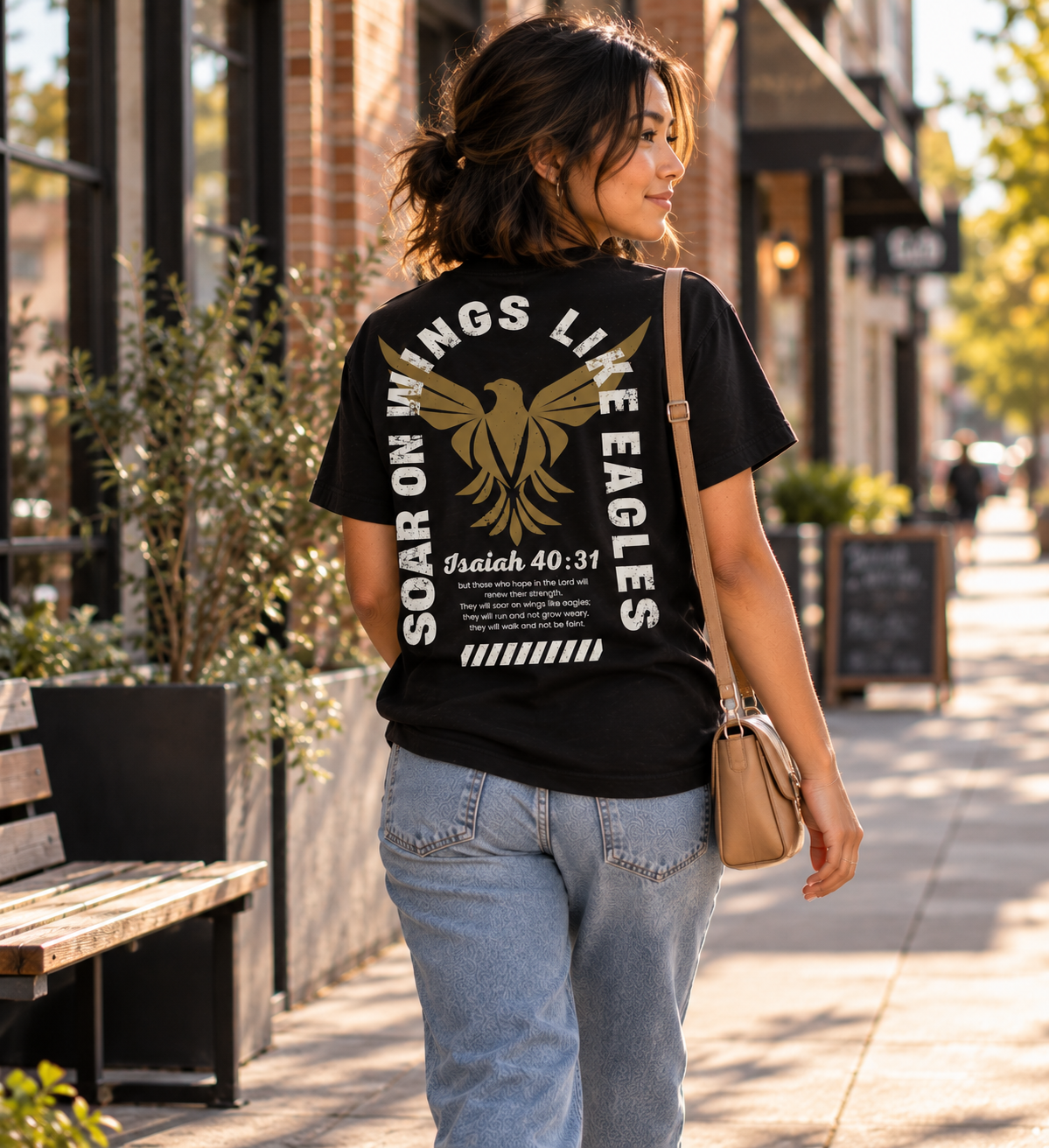 A woman walking on a city sidewalk viewed from behind, wearing a black graphic T-shirt with a large gold eagle and white text reading 'SOAR ON WINGS LIKE EAGLES' and 'Isaiah 40:31'.