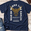 A navy blue short-sleeve t-shirt laid flat on a light wood surface, showing the back graphic with a large gold eagle, distressed white text reading 'Soar on Wings Like Eagles' and 'Isaiah 40:31', plus smaller scripture text. Jeans and part of a white shoe are visible as styling props.